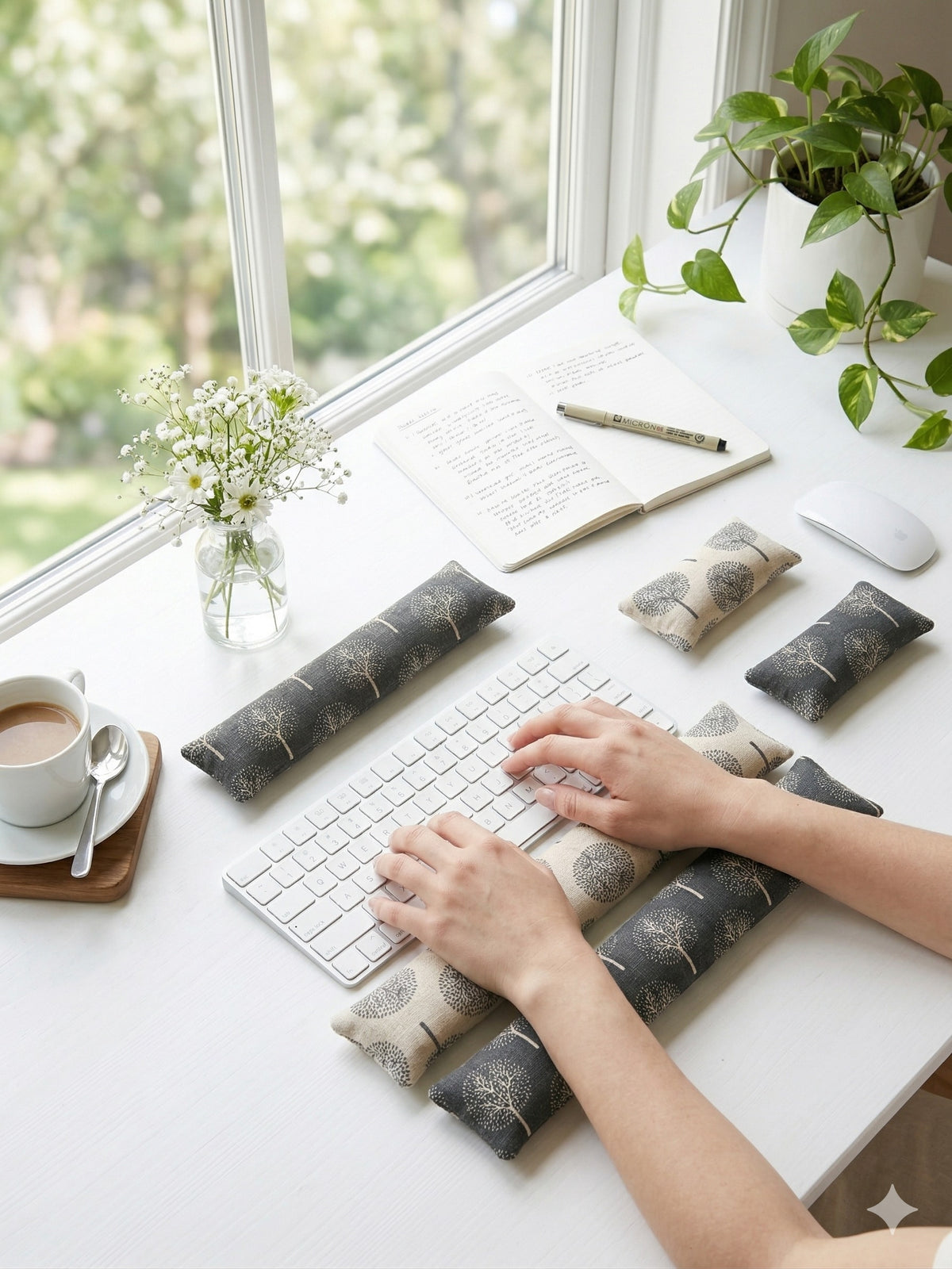 Hands typing on a white keyboard above tree print wrist rests on a bright desk by the window
